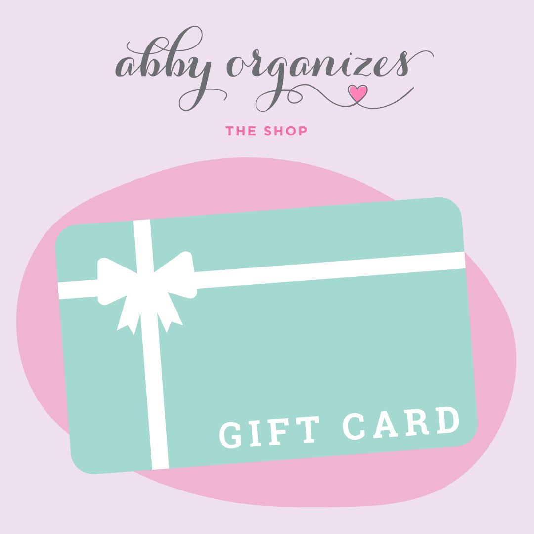 Illustration of a teal Abby Organizes Gift Card featuring a white ribbon design against a pink background. The text reads "Abby Organizes" at the top and "GIFT CARD" on the card, ideal for those who appreciate organization and thoughtful gifting.