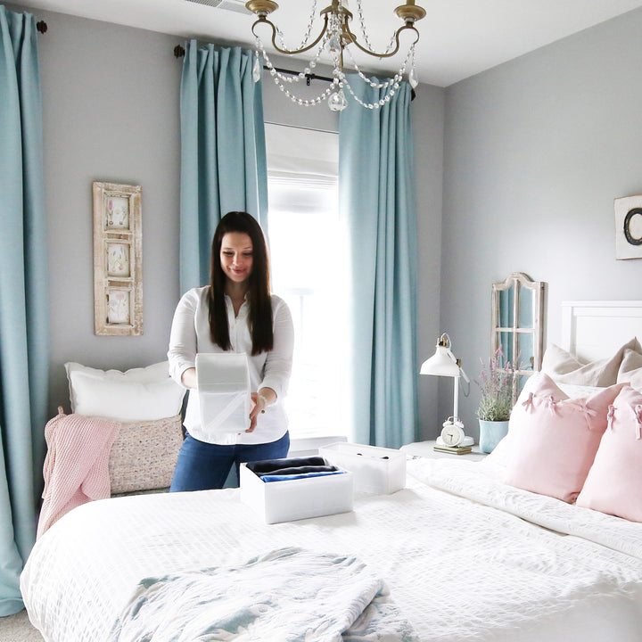 Woman organizing clothes into boxes on a bed, in a room with aqua curtains