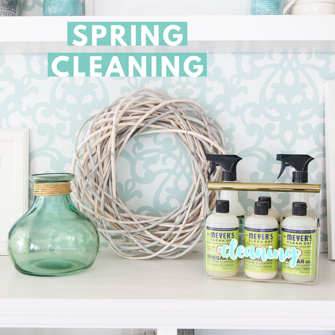 A decorative spring cleaning setup with a glass jar, a white wreath, and Mrs. Meyer’s cleaning products on a shelf, with “Spring Cleaning” text above on a light blue patterned background.
